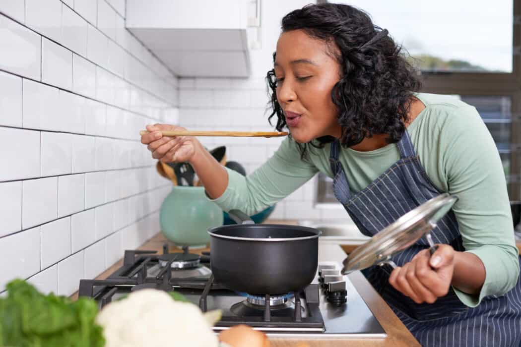 A young woman smells and tastes her cooking on the stove